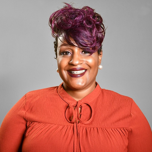 Portrait of a Black Lady Smiling at the Camera wearing a red jumper with a grey backdrop. Chanda