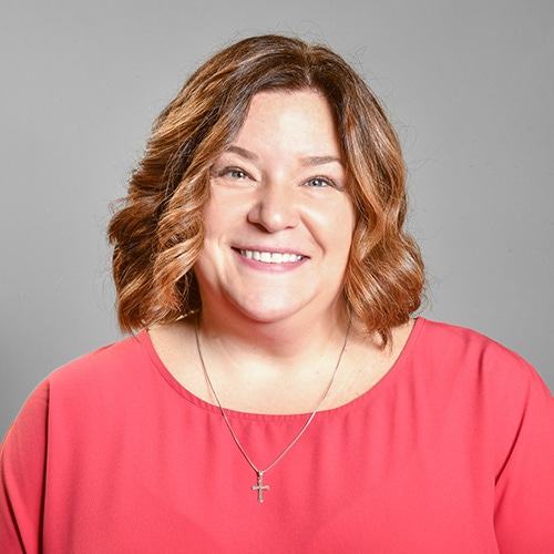 Portrait of a White Lady Smiling at the Camera wearing a red T-Shirt with a grey backdrop. Mary