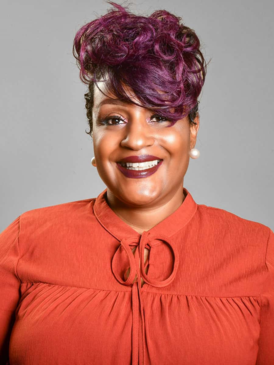 Portrait of a Black Lady Smiling at the Camera wearing a red jumper with a grey backdrop. Chanda