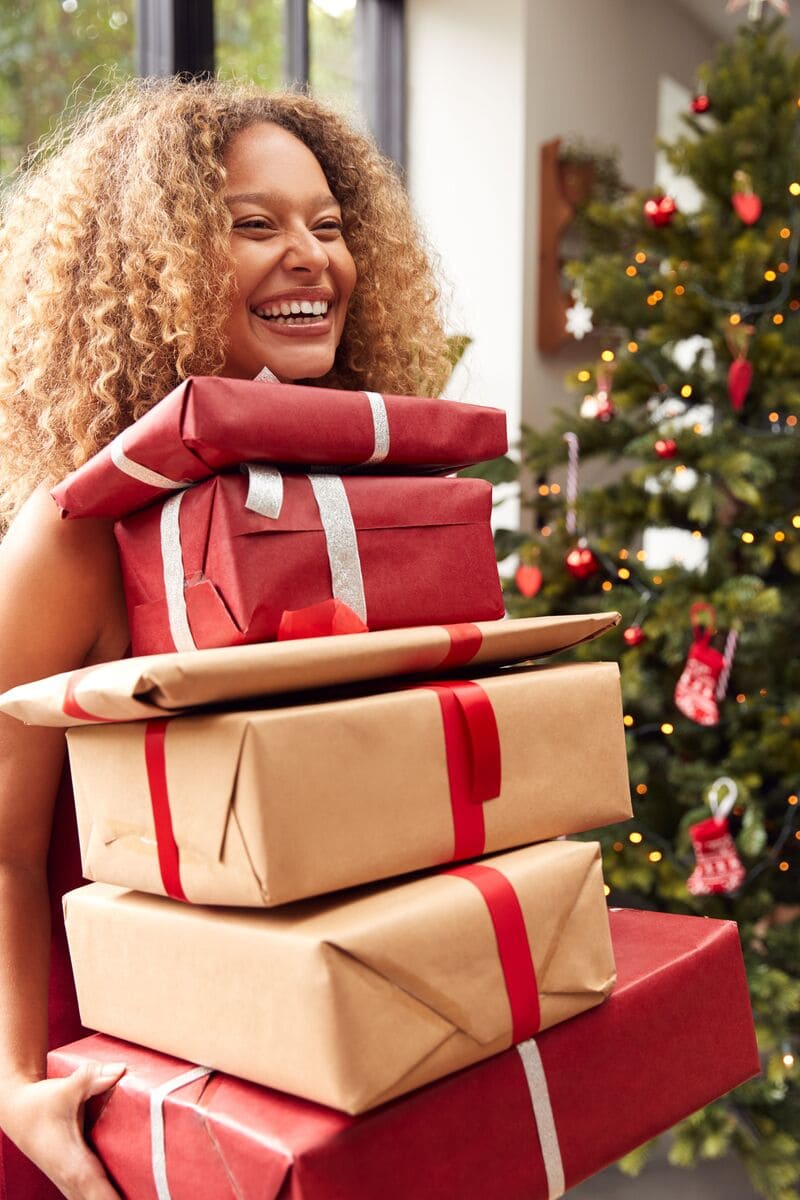 Woman Carrying Pile Of Christmas Presents Standing By Tree At Home
